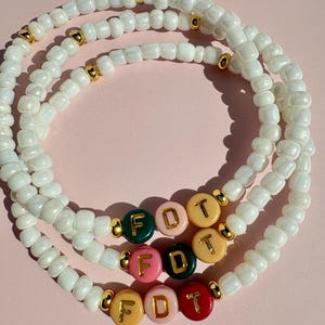 May include: Three beaded bracelets with white beads and gold accents. Each bracelet has a section with colorful letter beads spelling out "FDT". The bracelets are arranged on a pink surface.