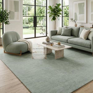 May include: A large, rectangular, sage green area rug with a textured surface covering a light wood floor. The rug is positioned in a living room featuring a matching sage green sofa, a rounded armchair, and a stone coffee table with books.