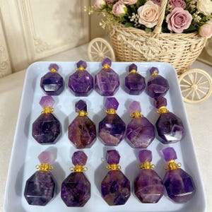 Peut inclure: Une collection de flacons de parfum en améthyste, avec des corps facettés et des bouchons géométriques. Les flacons sont d'un violet profond avec des accents dorés. Ils sont disposés sur un plateau blanc, avec un panier de fleurs en arrière-plan.