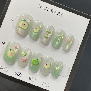 May include: A set of artificial nails with a pearlescent finish and apple-themed designs. The nails feature green and pink apple slices, whole apples, and small star and butterfly accents. The card reads "NAIL & ART".