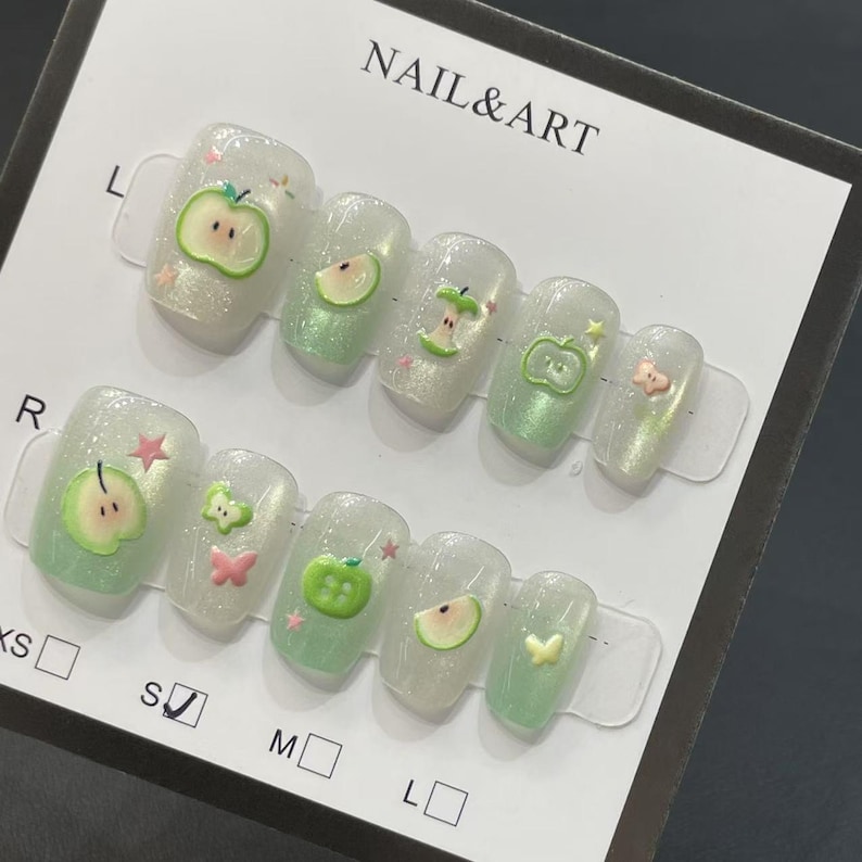 May include: A set of artificial nails with a pearlescent finish and apple-themed designs. The nails feature green and pink apple slices, whole apples, and small star and butterfly accents. The card reads "NAIL & ART".