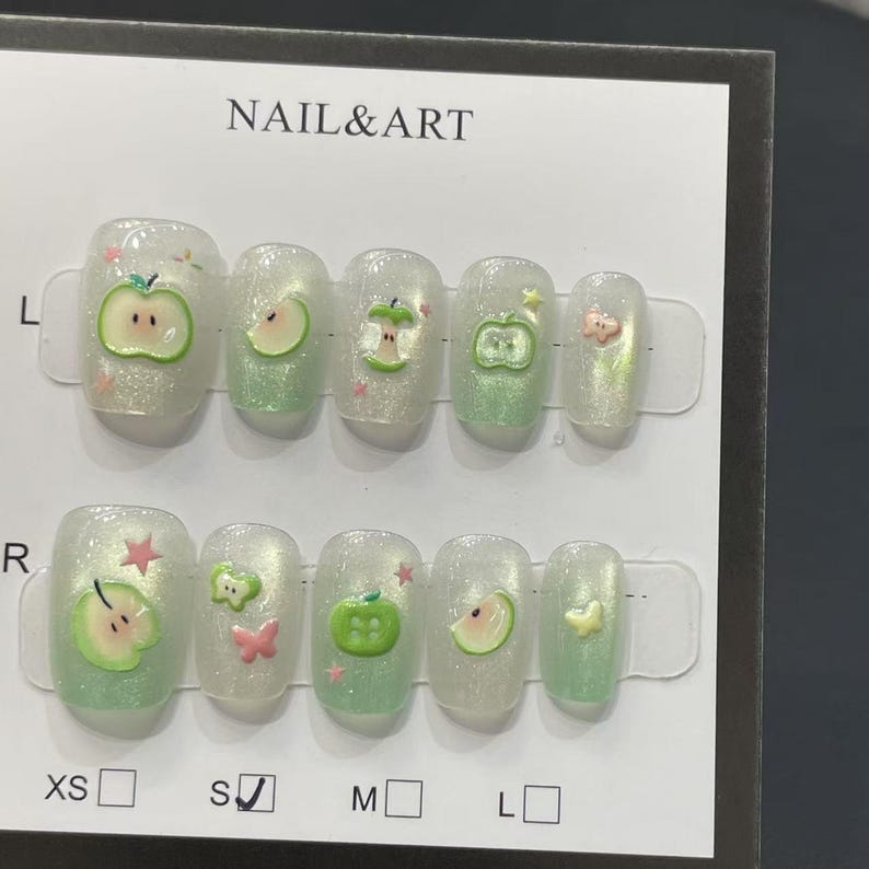 May include: A set of press-on nails with a clear, glittery base. Each nail features a green apple design with pink accents. The text "NAIL&ART" is visible at the top of the packaging. Sizes XS, S, M, and L are indicated.