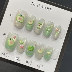May include: A set of press-on nails with a clear, glittery base and apple-themed designs. Each nail features a different apple design, including whole apples, slices, and a butterfly accent. The text "NAIL & ART" is visible.