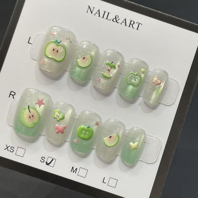 May include: A set of press-on nails with a clear, glittery base and apple-themed designs. Each nail features a different apple design, including whole apples, slices, and a butterfly accent. The text "NAIL & ART" is visible.