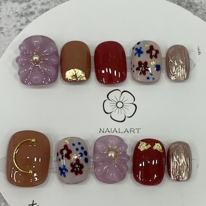 Puede incluir: Un conjunto de diez uñas artificiales con varios diseños. Las uñas presentan colores como malva, marrón y rojo, con detalles de pan de oro y motivos florales. Algunas uñas tienen una perla. La marca "NAIALART" está impresa en el embalaje.