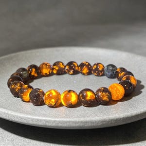 Könnte beinhalten: Ein Armband mit runden, bernsteinfarbenen Perlen mit dunkelbraunen und orangefarbenen Einschlüssen. Das Armband wird auf einem grauen Teller präsentiert, der die natürlichen Variationen der Edelsteinperlen hervorhebt. Es ist ein Schmuckstück.