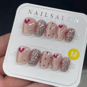 May include: A set of medium-sized, press-on nails with a pink base. Some nails feature red hearts, glitter, and small rhinestones. The package has the text "NAIL SALON" and "M" for size.