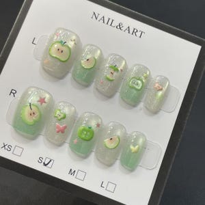 May include: A set of artificial nails with a pearlescent finish, decorated with green apple and pink butterfly designs. The nails are arranged on a white card with the text "NAIL & ART". Sizes are indicated with letters.