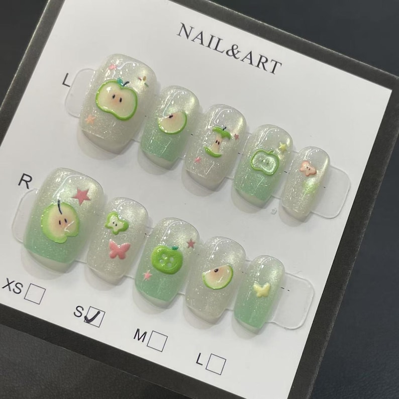 May include: A set of artificial nails with a pearlescent finish, decorated with green apple and pink butterfly designs. The nails are arranged on a white card with the text "NAIL & ART". Sizes are indicated with letters.