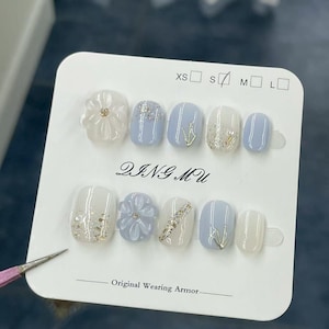 Puede incluir: Un conjunto de uñas postizas con varios diseños y tamaños. Las uñas presentan una mezcla de colores blanco y azul claro, con detalles florales y brillantes. Se puede ver el texto "DING MU" y "Original Wearing Armor".