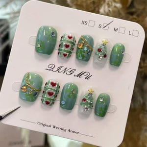 Könnte beinhalten: Ein Set künstlicher Fingernägel in verschiedenen Designs. Einige Nägel sind hellgrün mit floralen Akzenten und Strasssteinen. Andere zeigen rote Herzen, goldene Ketten und ein Weihnachtsbaum-Design. Der Text "DINGMU" und "Original Wearing Armor" sind sichtbar.