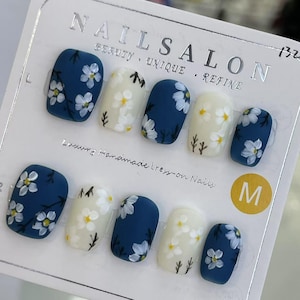 Puede incluir: Un juego de uñas postizas con acabado mate. Las uñas alternan entre azul oscuro y blanco roto, cada una adornada con delicados diseños florales blancos y tallos verdes. El paquete incluye el texto "NAILSALON" y "Luxury Handmade Press-on Nails."