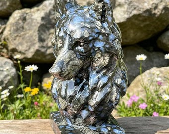 Puede incluir: Una escultura detallada de la cabeza de un lobo, hecha de piedra con un patrón moteado de negro, gris y azul. El lobo se representa en una vista de tres cuartos, con la cabeza y los hombros visibles. La escultura se asienta sobre una base rectangular.