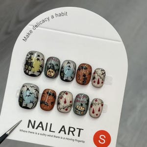 May include: A set of Halloween-themed press-on nails. Each nail features a different design, including haunted houses, pumpkins, and bats. The nails are displayed on a white card with the text "NAIL ART". The card also has the text "Make delicacy a habit".