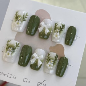 Puede incluir: Un conjunto de uñas postizas con varios diseños. Algunas uñas son de color verde oliva liso, otras tienen bases blancas con detalles florales y de hojas. Dos uñas presentan detalles de lazo blanco. Las uñas se muestran en una tarjeta blanca.