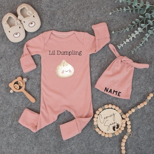 May include: A baby gift set in a soft pink color, including a ribbed jumpsuit with "Lil Dumpling" text and a dumpling graphic, a matching knotted hat with "NAME" text, and a wooden rattle. Also included are a wooden sign with "Coming Soon" text and baby feet, and a pair of bear-faced baby shoes.