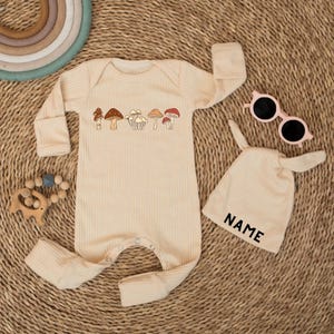 May include: Cream-colored baby romper and matching hat set. The romper features a row of illustrated mushrooms and long sleeves. The hat has bunny ears and the word "NAME" printed on it. Pink sunglasses are also included.
