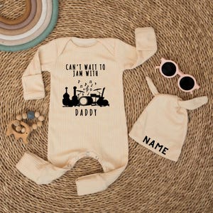 May include: A cream-colored baby onesie with the text "CAN'T WAIT TO JAM WITH DADDY" and a musical instrument graphic. Includes a matching hat with "NAME" printed on it and pink sunglasses. A wooden teething toy and a rainbow are also visible.