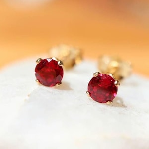 May include: A pair of stud earrings featuring round, faceted, deep red gemstones set in gold-toned metal prongs. The earrings are positioned on a textured, off-white surface, showcasing the brilliant cut and vibrant colour of the stones.