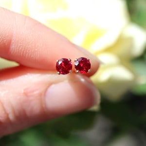 May include: A pair of stud earrings with bright red gemstones set in gold. The round-cut stones are a deep red colour, and the earrings are held up by a hand against a blurred background of yellow and green.