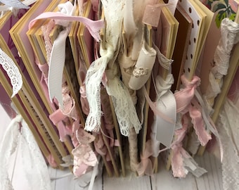 May include: A handmade scrapbook with pink and cream-colored pages. The pages are bound together with various ribbons and lace in shades of white, cream, and pink. The scrapbook has a decorative, feminine aesthetic.