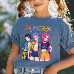 May include: A blue t-shirt featuring the word "HUNTRAX" in colorful text above a graphic of three anime-style girls. The girls are wearing different outfits, including shorts and jackets. The girl in the middle is wearing a yellow jacket.
