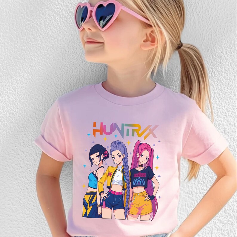 May include: A light pink t-shirt featuring a colorful graphic of three anime-style girls. The word "HUNTRAX" is printed above the graphic. The girls are wearing different outfits, including crop tops, shorts, and jackets.