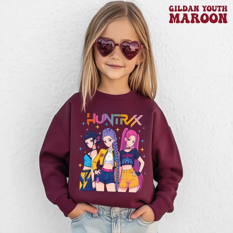 May include: Maroon Gildan Youth sweatshirt featuring a colorful graphic of three anime-style girls. The word "HUNTRX" is printed above the graphic. The girl is wearing heart-shaped sunglasses.