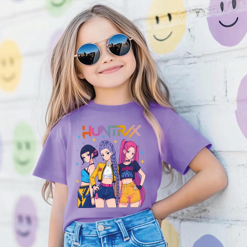 May include: A purple t-shirt featuring the word "HUNTRAX" in colorful text above an anime-style illustration of three girls. The girls are wearing different outfits, including shorts and crop tops. The background is a white brick wall with colorful smiley face designs.