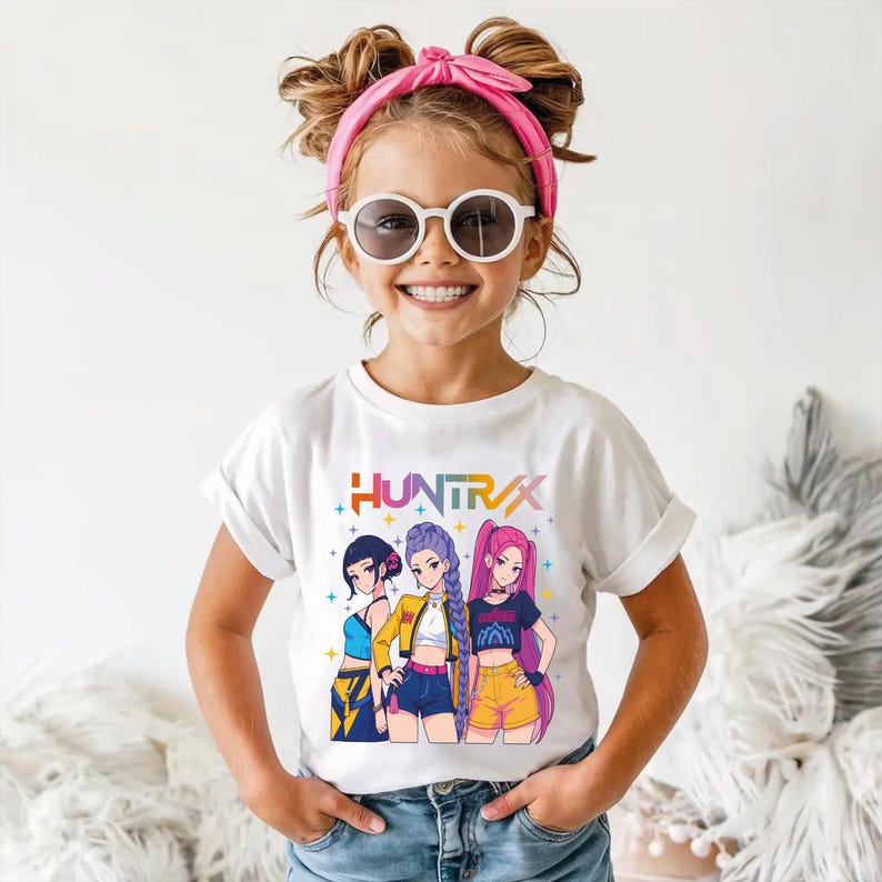 May include: White t-shirt featuring the word "HUNTRAX" in colorful text above a graphic of three anime-style girls. The girls are wearing different outfits, including shorts and crop tops. The girl in the middle is wearing a yellow jacket.