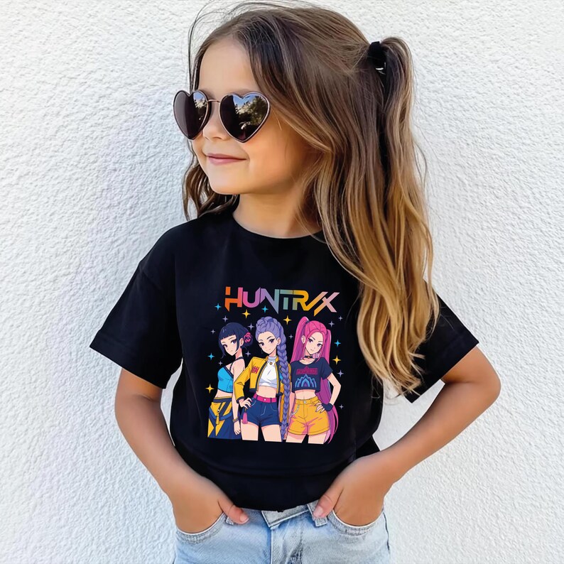May include: A black t-shirt featuring a colorful graphic of three anime-style girls with the word "HUNTRAX" in a rainbow font. The girls are wearing different outfits, including shorts and jackets. The girl in the center is wearing a yellow jacket.