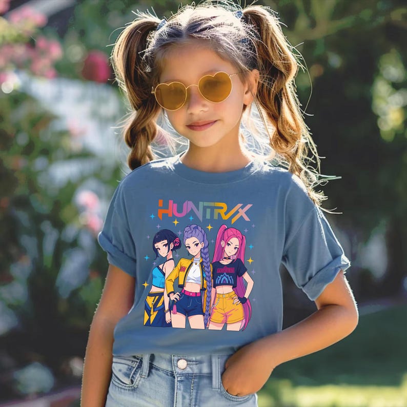 May include: A blue t-shirt featuring the word "HUNTRAX" in colorful text above a graphic of three anime-style girls. The girls are wearing different outfits, including shorts and jackets. The girl in the middle is wearing a yellow jacket.