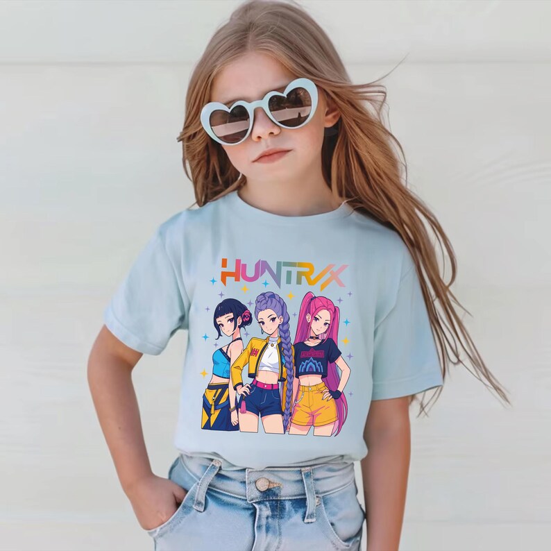 May include: A light blue t-shirt featuring a colorful graphic of three anime-style girls with the text "HUNTRX". The girls are wearing different outfits, including shorts, tops, and jackets. The girl in the middle is wearing a yellow jacket.
