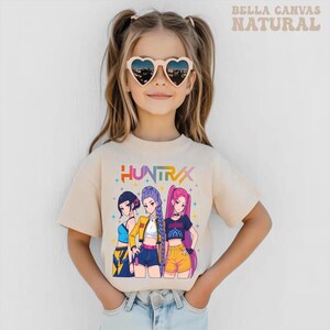May include: A beige t-shirt featuring the word "HUNTRAX" above a graphic of three anime-style girls. The girls are wearing colorful outfits. The shirt is worn by a young person wearing heart-shaped sunglasses.
