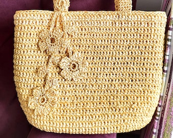 May include: A light yellow, hand-crocheted tote bag featuring a textured, woven pattern. The bag has two sturdy top handles and is decorated with three matching crocheted flower embellishments attached to the front side by a thin cord.