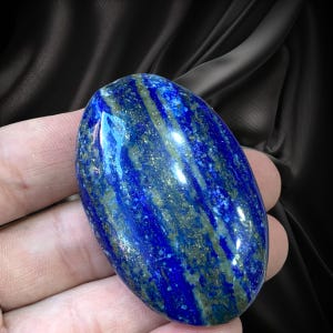 May include: An oval-shaped lapis lazuli gemstone featuring a deep royal blue base color with prominent streaks of golden pyrite inclusions and subtle greyish-white mineral veins throughout the polished, smooth, and reflective surface of the stone.