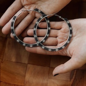 May include: A pair of large hoop earrings with a beaded design. The earrings feature a silver metal base and are adorned with black and white beads in a repeating pattern. The earrings are held in a person's hands.