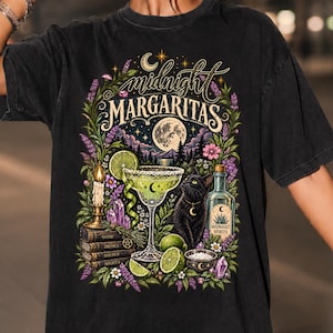 以下が含まれることがあります： 「midnight MARGARITAS」の文字がプリントされた黒の半袖Tシャツ。デザインには、マルガリータグラス、黒猫、「MIDNIGHT SPIRITS」と書かれたボトル、積み重ねられた本、キャンドル、花などのイラストが描かれています。
