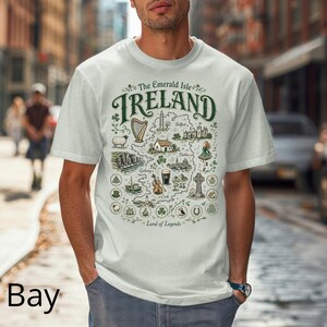 Puede incluir: Una camiseta de color claro con un mapa gr&aacute;fico detallado de Irlanda. El dise&ntilde;o incluye el texto "The Emerald Isle IRELAND" y "Land of Legends", junto con ilustraciones de un arpa, ovejas, acantilados, una pinta de cerveza y un viol&iacute;n.
