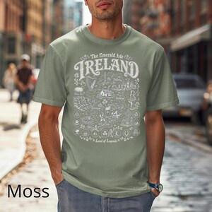 Puede incluir: Una camiseta de manga corta color verde musgo con un estampado gr&aacute;fico blanco de un mapa de Irlanda. El texto dice The Emerald Isle IRELAND Land of Legends, acompa&ntilde;ado de ilustraciones de un arpa, ovejas, acantilados y varios s&iacute;mbolos.