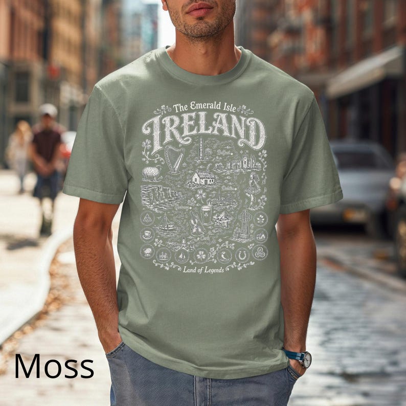 Puede incluir: Una camiseta de manga corta color verde musgo con un estampado gr&aacute;fico blanco de un mapa de Irlanda. El texto dice The Emerald Isle IRELAND Land of Legends, acompa&ntilde;ado de ilustraciones de un arpa, ovejas, acantilados y varios s&iacute;mbolos.