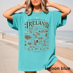Puede incluir: Una camiseta de manga corta color azul cerceta con un estampado gr&aacute;fico de un mapa de Irlanda. El dise&ntilde;o incluye el texto "The Emerald Isle IRELAND Land of Legends" junto con ilustraciones de un arpa, ovejas, acantilados, una pinta y una cruz.