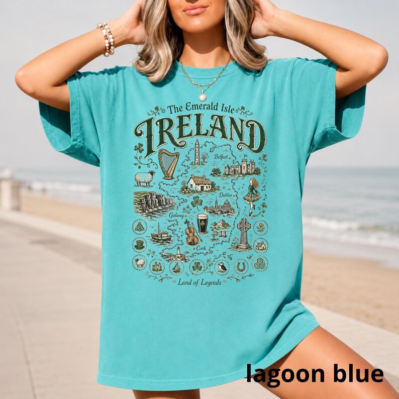 Puede incluir: Una camiseta de manga corta color azul cerceta con un estampado gr&aacute;fico de un mapa de Irlanda. El dise&ntilde;o incluye el texto "The Emerald Isle IRELAND Land of Legends" junto con ilustraciones de un arpa, ovejas, acantilados, una pinta y una cruz.
