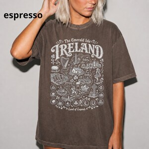 Puede incluir: Una camiseta de manga corta color espresso con un estampado gr&aacute;fico blanco de un mapa de Irlanda. El dise&ntilde;o incluye el texto The Emerald Isle, IRELAND y Land of Legends, rodeado de ilustraciones de monumentos, s&iacute;mbolos y motivos florales.
