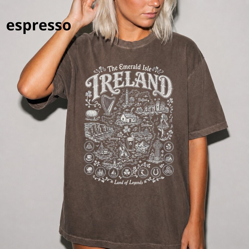 Puede incluir: Una camiseta de manga corta color espresso con un estampado gr&aacute;fico blanco de un mapa de Irlanda. El dise&ntilde;o incluye el texto The Emerald Isle, IRELAND y Land of Legends, rodeado de ilustraciones de monumentos, s&iacute;mbolos y motivos florales.