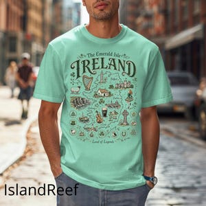 Puede incluir: Una camiseta de color verde menta claro con un mapa gr&aacute;fico detallado de Irlanda. El dise&ntilde;o incluye el texto The Emerald Isle IRELAND Land of Legends, junto con ilustraciones de un arpa, ovejas, acantilados, un viol&iacute;n y una cruz celta.
