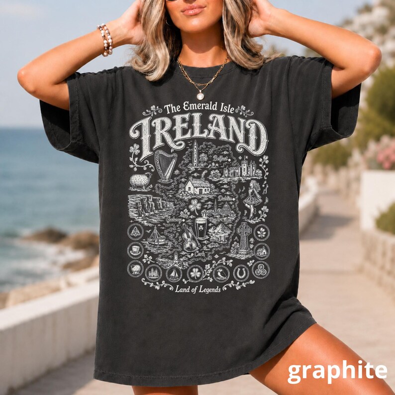 Puede incluir: Una camiseta de manga corta gris carb&oacute;n con un estampado gr&aacute;fico blanco de un mapa de Irlanda. El dise&ntilde;o incluye ilustraciones de un arpa, un faro, una oveja, una pinta de cerveza y una cruz celta, con el texto The Emerald Isle IRELAND Land of Legends.
