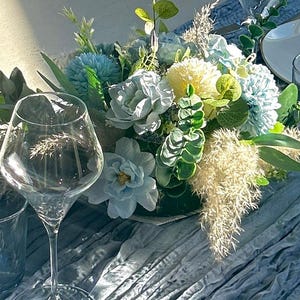 Peut inclure: Une table joliment dressée avec un centre de table floral composé de fleurs bleues et blanches, de feuilles vertes et d'éléments beiges. Un verre à vin et un gobelet bleu sont au premier plan. Une assiette blanche avec un bord argenté est visible.
