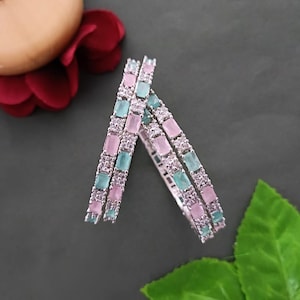 May include: Three silver bangles with alternating pastel pink and teal rectangular stones. The bangles are set against a dark gray background with a red flower and green leaf.