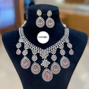 Può includere: Un set di gioielli abbagliante con collana e orecchini abbinati. La collana e gli orecchini sono adornati con cristalli trasparenti e gemme rosa a forma di goccia. Il set è esposto su una testa di manichino nera, mostrando il design.